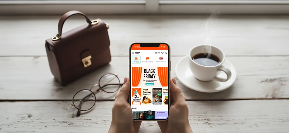A person holding a smartphone with the Kasta app open on the Black Friday page, next to a cup of coffee, glasses, and a handbag on a wooden table.