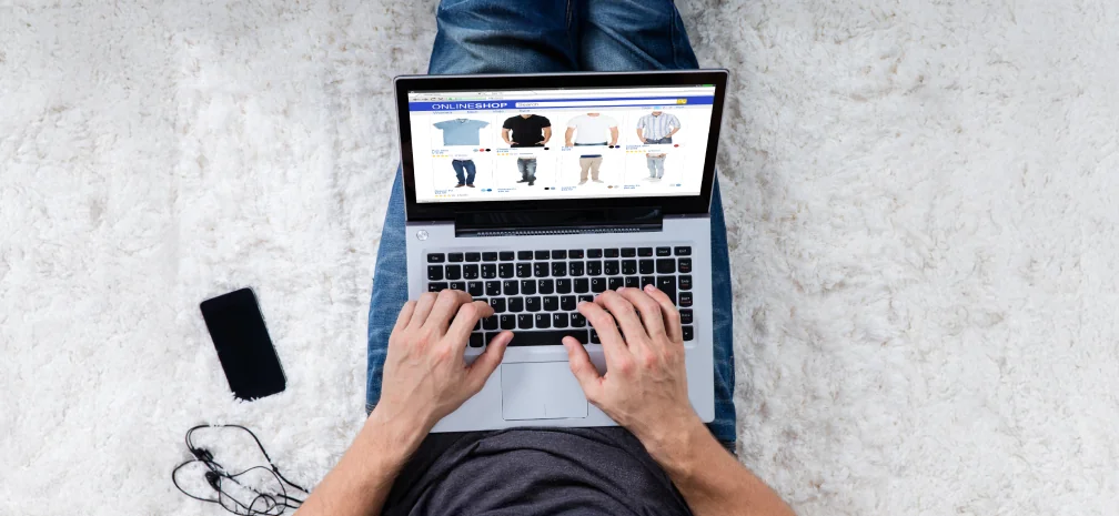 Top-down view of a person using a laptop to browse Google Shopping product listings, with a phone and earphones placed nearby.